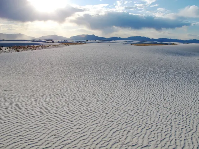 White dunes in foreground with sun setting behind mountain.