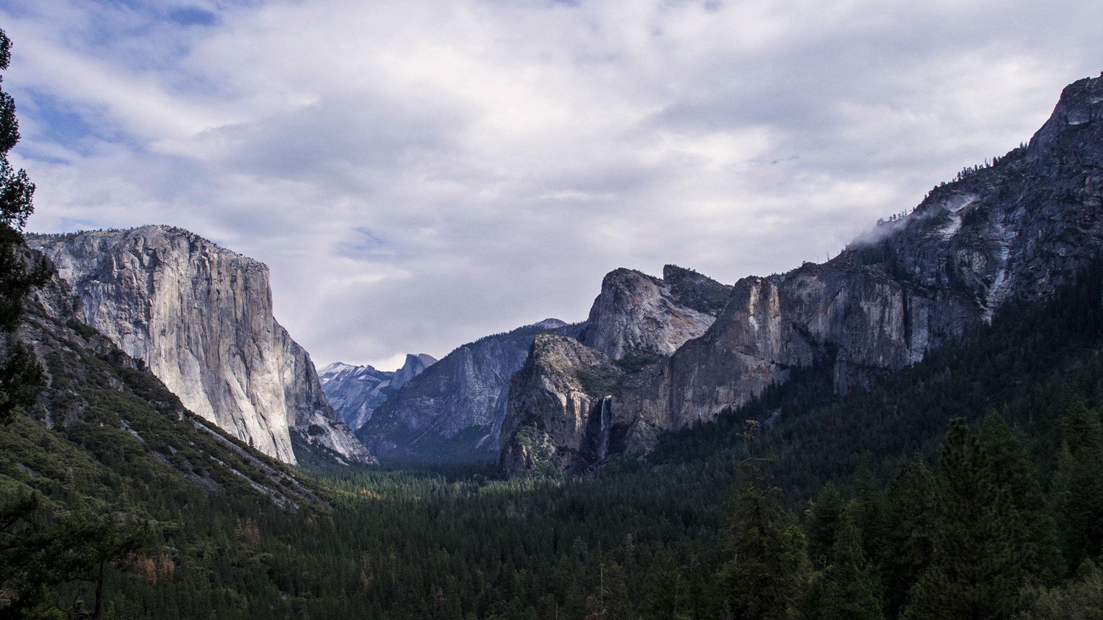 Granite cliffs and forested Yosemite Valley beneath a cloudy sky.